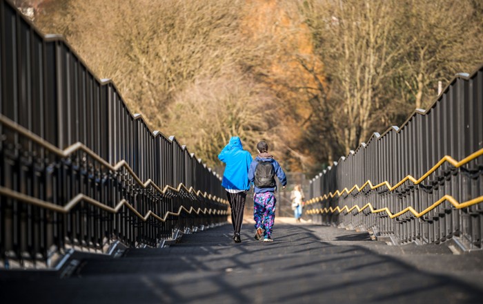 Two young people walk side by side down a wide pedestrian bridge or overpass lined with railings.