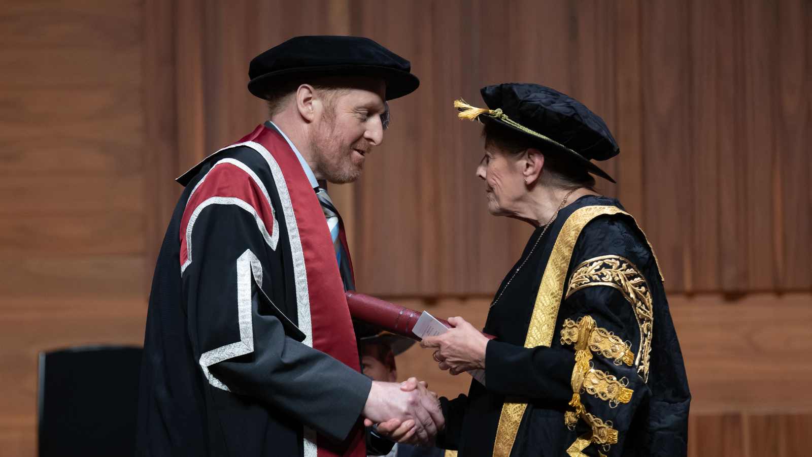 Man wearing cap & gown and holding a scroll shaking hands with the university's chancellor on stage