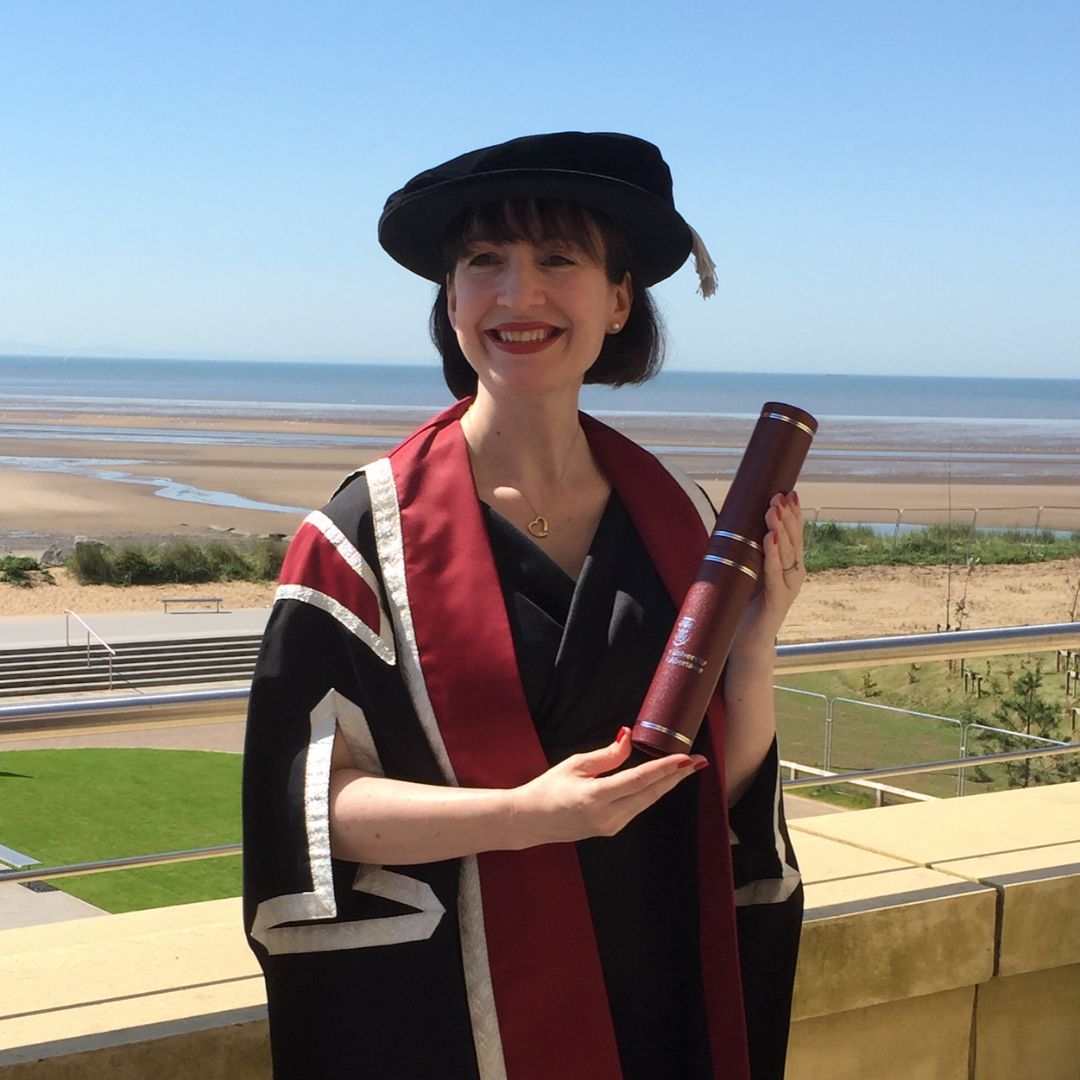 Lady wearing a cap and gown, holding a scroll with a beach backdrop, smiling at the camera