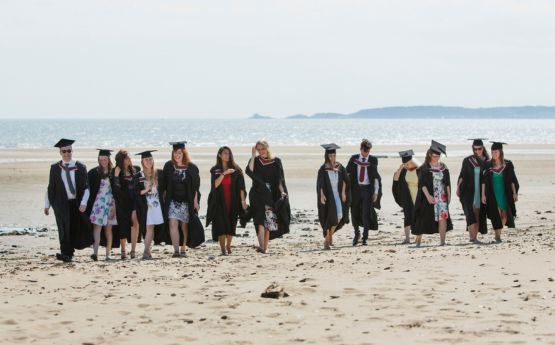 Students walking on the beach wearing cap and gown on graduation day with Mumbles pier in the background