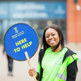 An image of a Student Ambassador holding a 'Here to Help' sign on the Bay Campus.