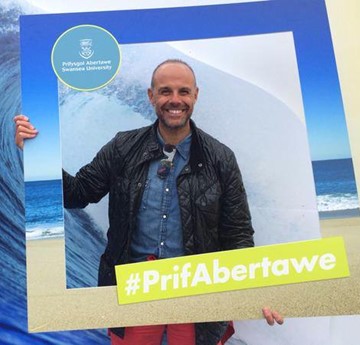 Jason Mohammad yn sefyll y tu ôl i ffrâm fawr sy’n dweud #PrifAbertawe