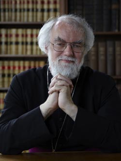 Llun o'r Gwir Barchedig yn eistedd mewn llyfrgell yn gwenu i'r camera