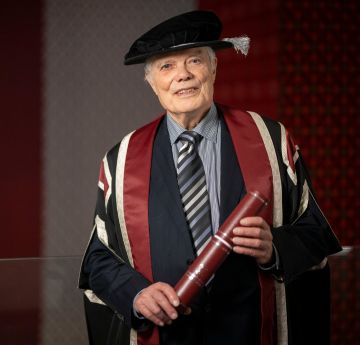 Dyn mewn gwisg graddio yn dal scroll yn gwenu wrth y camera