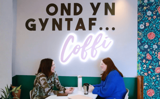 Dau person yn eistedd i cael coffi gyda arwydd ar y wal yn dweud