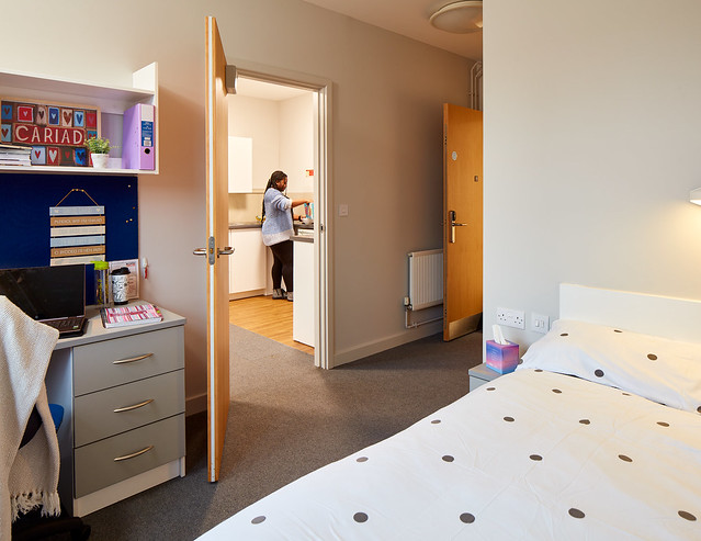 Student standing in kitchen with the bedroom in view in the foreground.