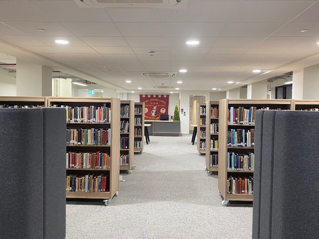 Golwg ar resi o silffoedd pren yn llawn llyfrau, ar ddwy ochr llwybr cerdded, yn edrych tuag at ardal y dderbynfa yn Llyfrgell Glowyr De Cymru. Mae baner goch o'r casgliad uwchben desg y dderbynfa.