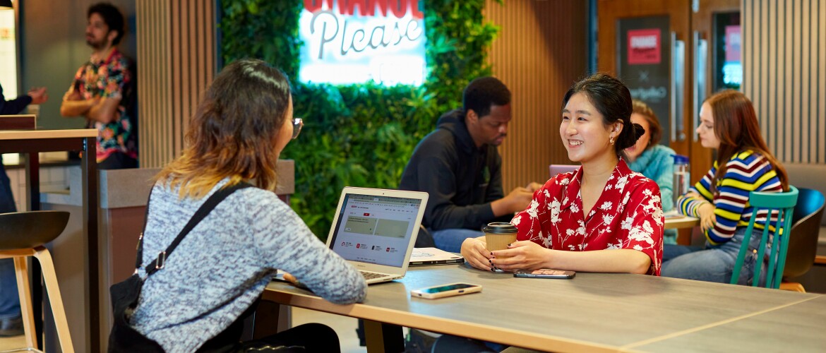 Two students sat at a table, one of which has a laptop