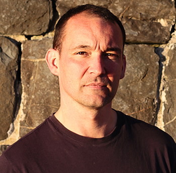 a photo of Cynan Jones from the chest up. He is wearing a dark sweater and is stood in front of a stone wall. Sunlight is casting a shadow across the right side of his face. 