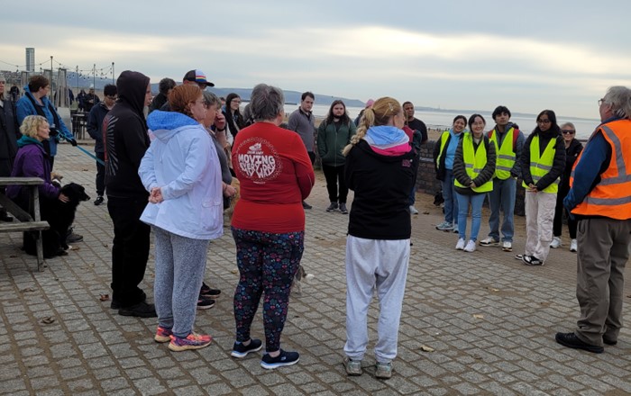 Grŵp o bobl yn sefyll y tu allan ar lan y môr yn gwrando ar ddyn mewn fest golygfa uchel