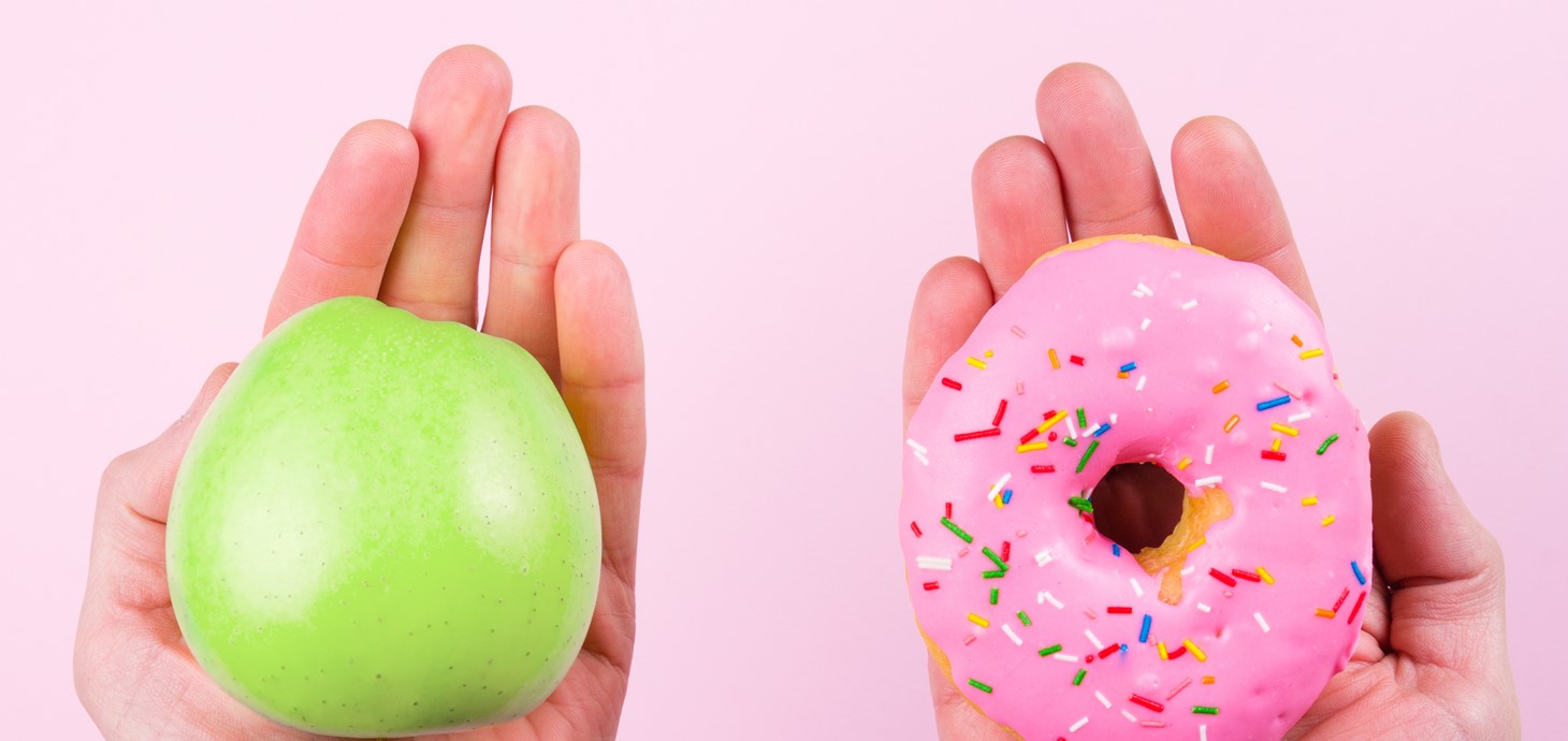 Two hands are holding items against a pink background: the left holds a green apple, and the right holds a pink donut with colorful sprinkles, suggesting a choice between healthy and indulgent.
