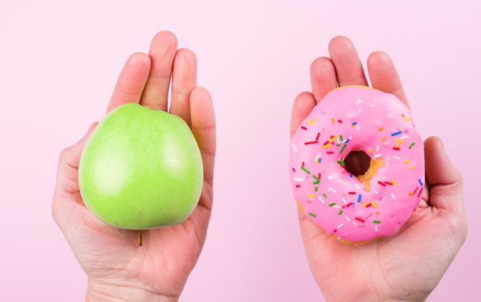 Two hands are holding items against a pink background: the left holds a green apple, and the right holds a pink donut with colorful sprinkles, suggesting a choice between healthy and indulgent.