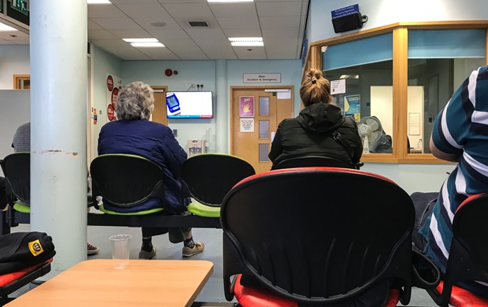 Pobl yn eistedd mewn ystafell aros ysbyty gyda ddesg dderbynfa yn y cornel