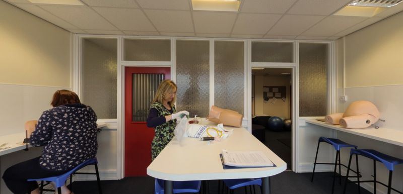 Lab sgiliau bydwreigiaeth gyda modelau hyfforddi a myfyrwyr yn ymarfer gofal