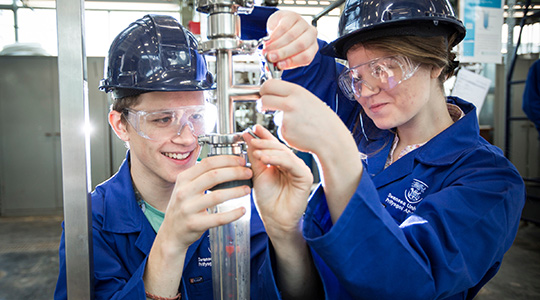 Two students wearing blue lab coats examine mechanical equipment