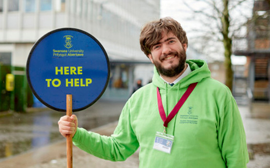 Student in lime green holding a 'Here to Help' lollipop sign