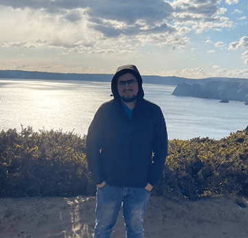 Our student Aser standing in a relaxed pose on the background of sea na cliffs, with sun rays shining on the sea surface.