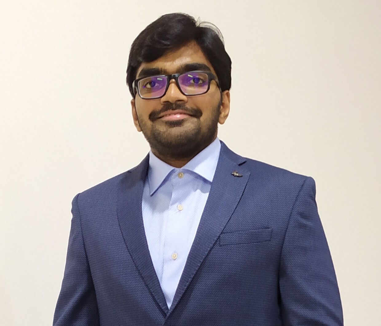 A photo of our graduate Sai in a smart navy suit smiling in the camera, on the cream background.