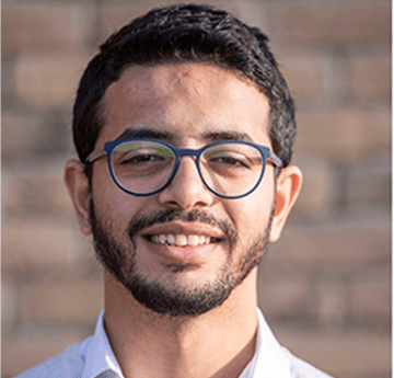 A portrait of Nabil, who is smiling at the camera with a light brick wall at the background.