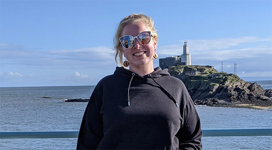 student standing in front of the sea, smiling towards the camera