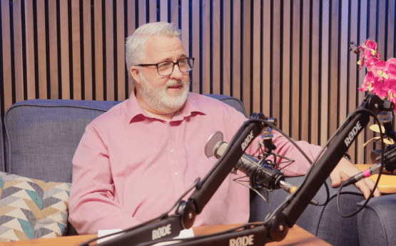 A man sitting on a grey sofa speaking into a microphone for a podcast.
