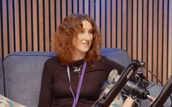 A woman sitting on a grey sofa speaking into a microphone for a podcast.