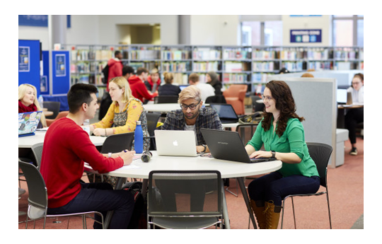Students sat around desks studying. They are using laptops and books. There are bookshelves in the background.