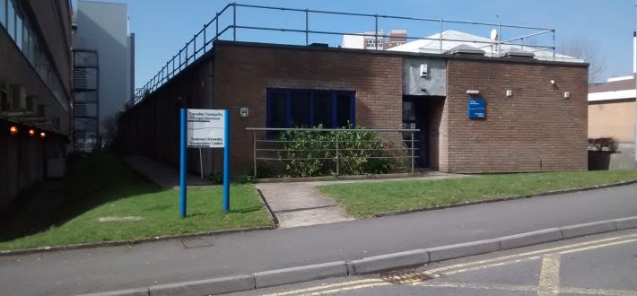 The image shows the front view of the Amy Dillwyn Building in which the Transcription Centre is located. It shows a pathway with a sign on the left leading up to the automatic front door.