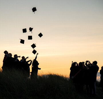 Students at graduation, throwing caps in the air as the sun sets.
