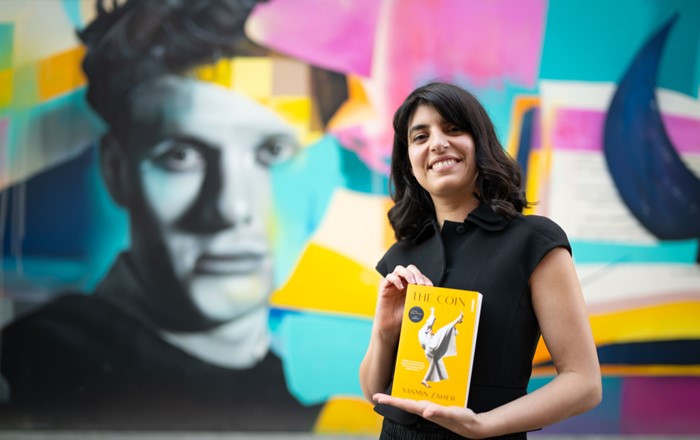 Yasmin Zaher stands holding her novel The Coin in front of a colourful mural of Dylan Thomas