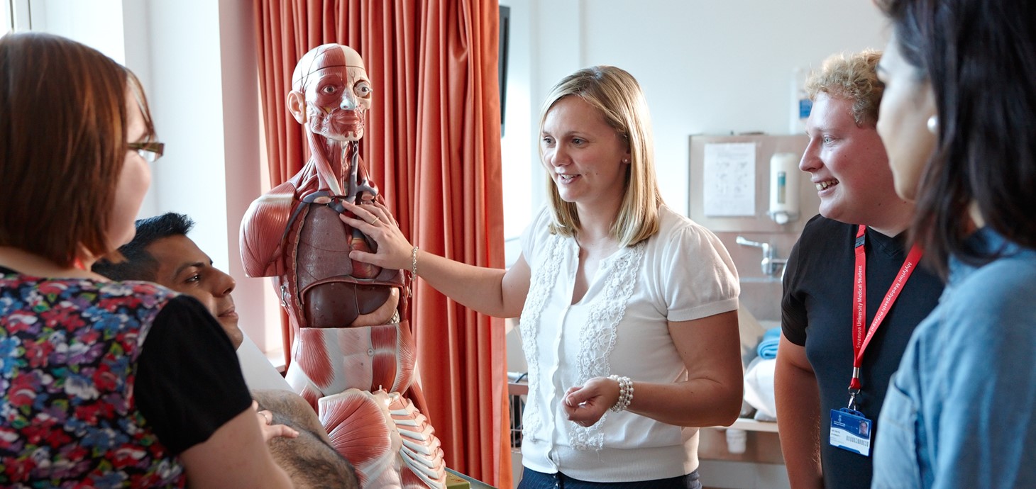 Group of students in medical practice class looking at a skeleton while listening to a lecturer