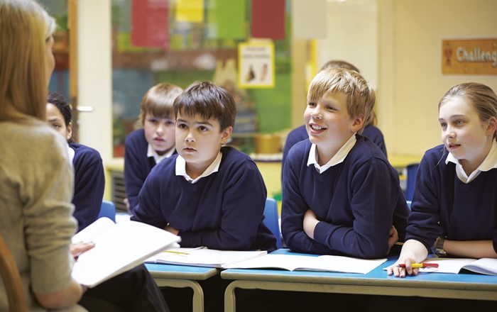 Teacher sitting facing a group of school pupils in uniform who are listening to her reading from a book on her lap. 