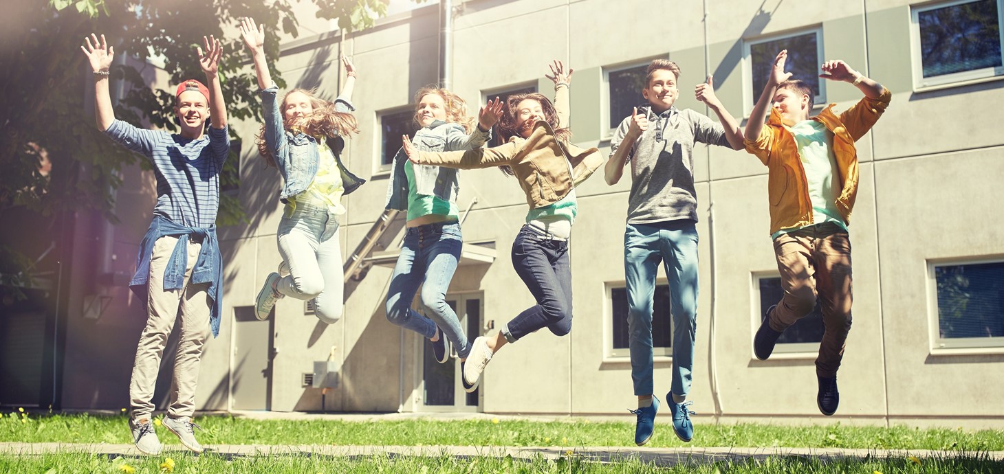 Group of six casually dressed teenagers - three boys and three girls, smiling and jumping in the air outside a building.