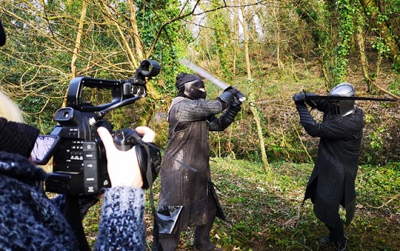 Two men dressed as medieval knights in armour fighting outdoors with swords while being photographed by a person with a camera. 