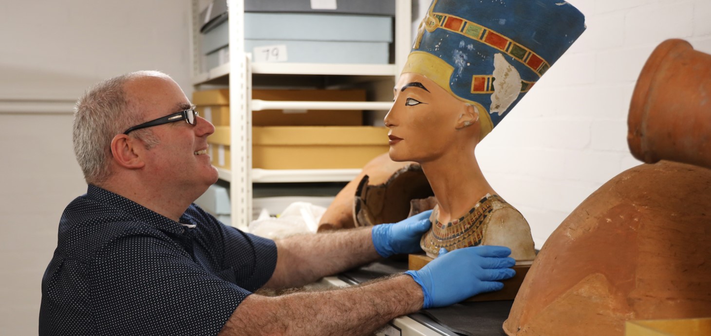 Man smiling inside a storeroom surrounded by shelves holding a variety of Egyptian artefacts.