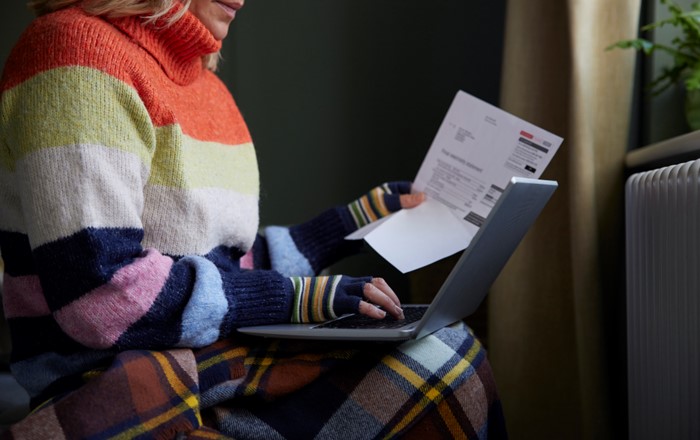 A woman wrapped in a blanket and wearing a thick jumper and gloves looks at a laptop while holding a utility bill in her hand.