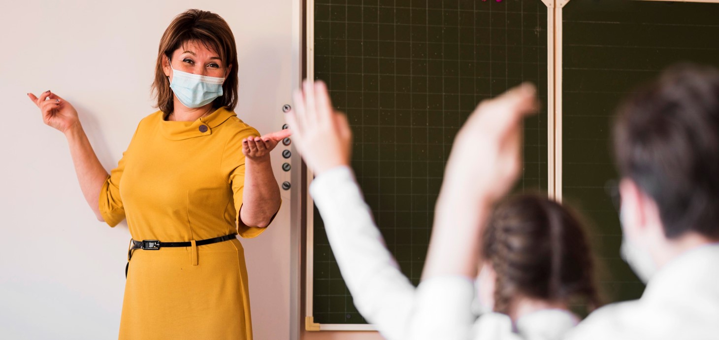 Female teacher wearing a facemask standing at a white board at the front of pupils.