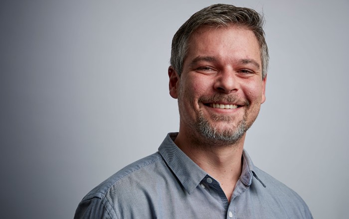 Head and shoulders photo of smiling man in open necked shirt against a grey background