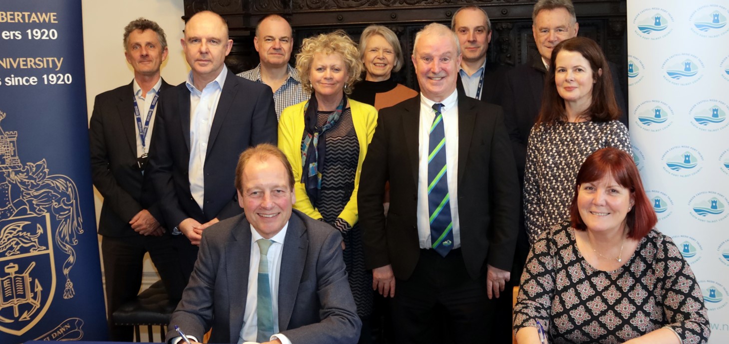 Man and woman sitting at table signing document with group of nine people standing behind them - all looking at the camera