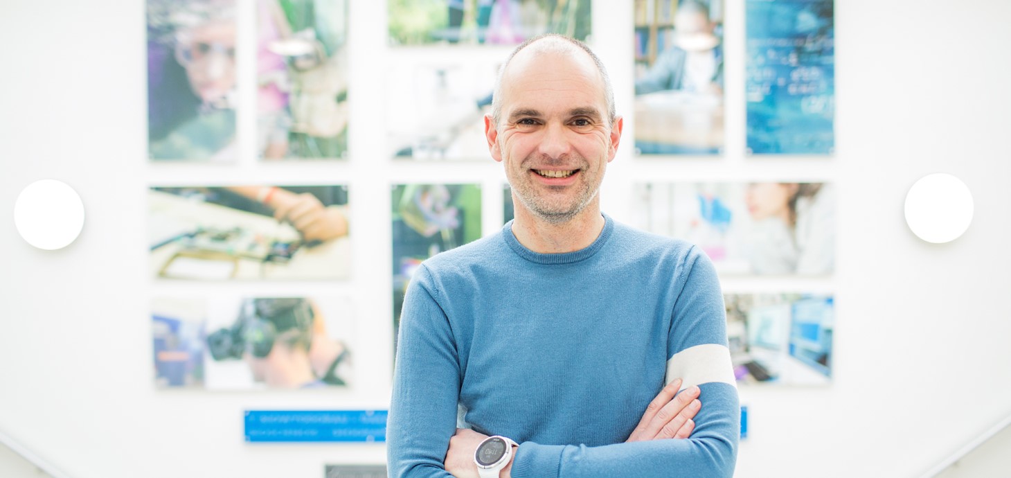 Professor Matt Jones is smiling and has his arms folded. He is wearing a blue sweater and standing in front of brightly coloured artwork.