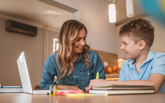 Woman sitting at table smiling at a boy sitting alongside her using a laptop