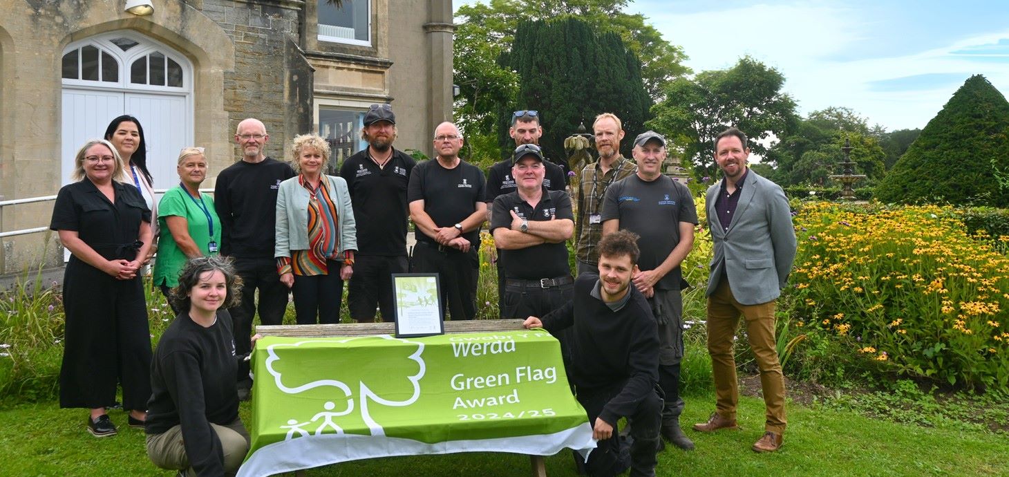 Group of people standing in a garden standing outside a building by a table draped with a green flag.