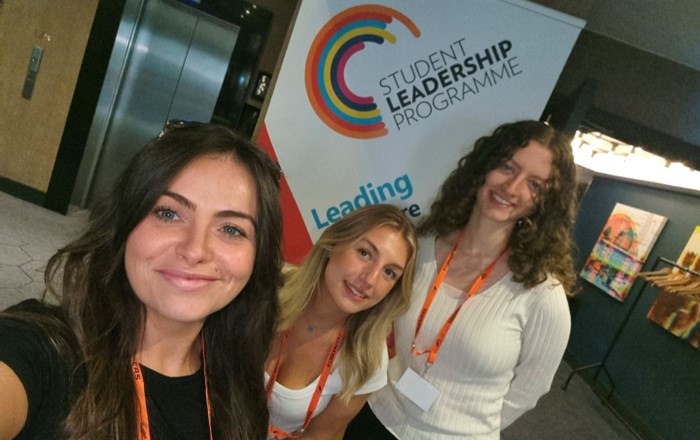 A selfie of Cerys, Lily, and Ellesse at a recent welcome event for the Student Leadership Programme. In the background is a pull-up banner with the programme’s logo on it.