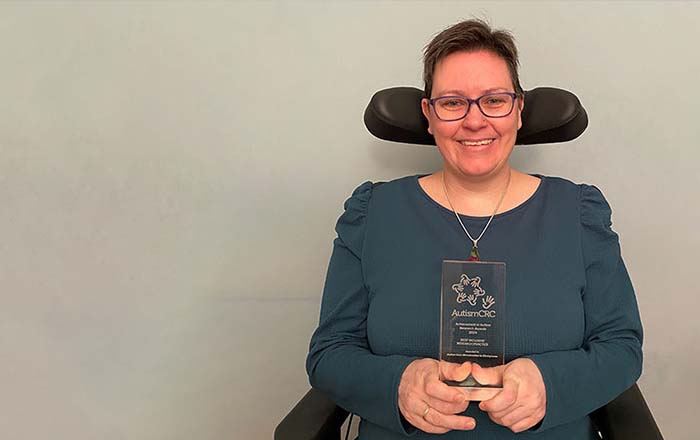 Alt-text Dr Aimee Grant is smiling as she holds her glass Autism CRC award. She is seated in her wheelchair. 