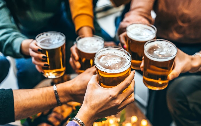 Shot of a group of people's hands. They are all holding full pint glasses of beer above a table top and clinking them together to raise a toast.