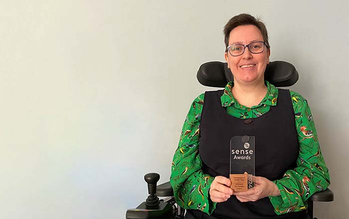 Dr Aimee Grant is smiling as she holds her glass Sense award. She is seated in her wheelchair and wears a black dress and green blouse.