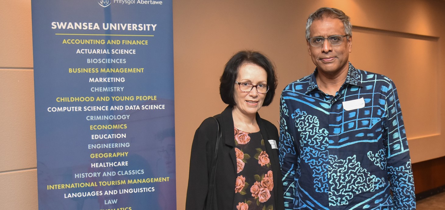 Man and a woman standing indoors next to a banner promoting Swansea University