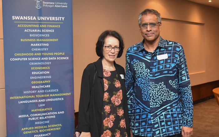 Man and a woman standing indoors next to a banner promoting Swansea University