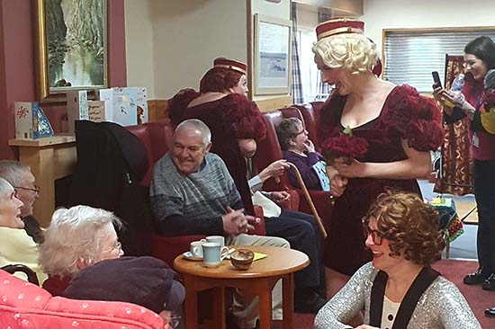 Group of elderly people sitting down in care home setting talking to women in costumes