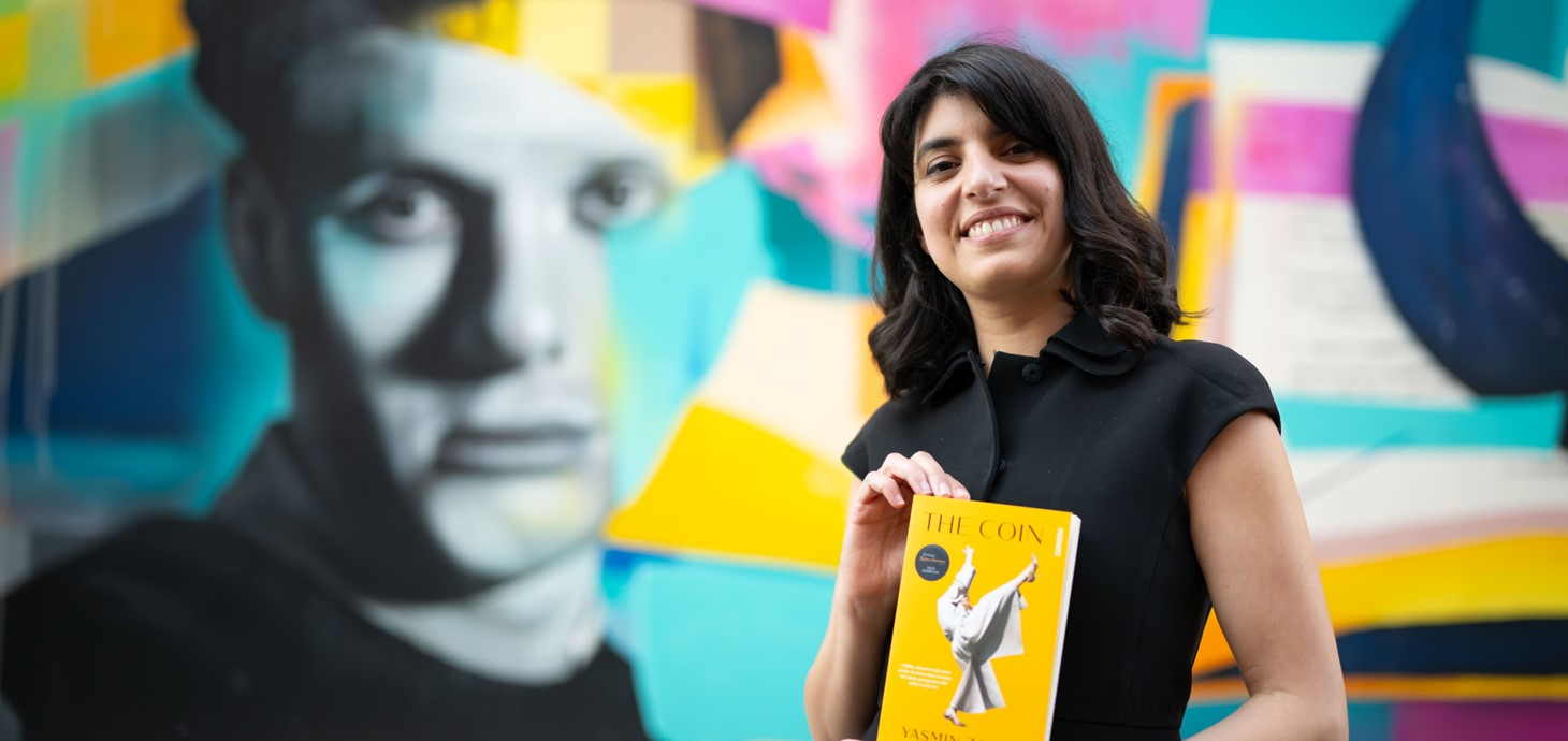 Yasmin Zaher stands holding a copy of her novel,The Coin, in front of a colourful mural featuring the face of Dylan Thomas.